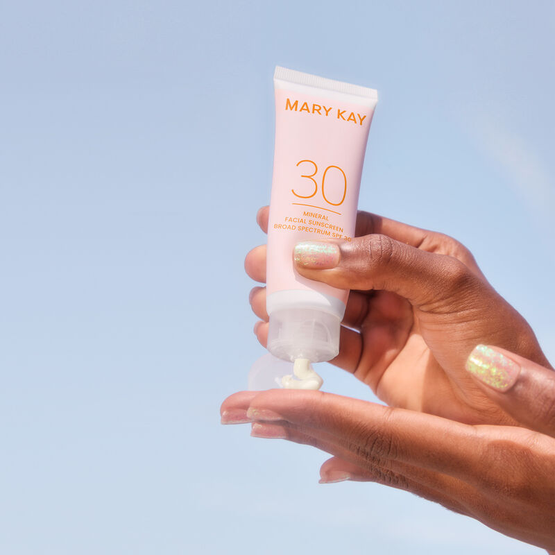 Deep-skinned hands squeezing tube of Mary Kay Mineral Facial Sunscreen SPF 30 against a blue sky