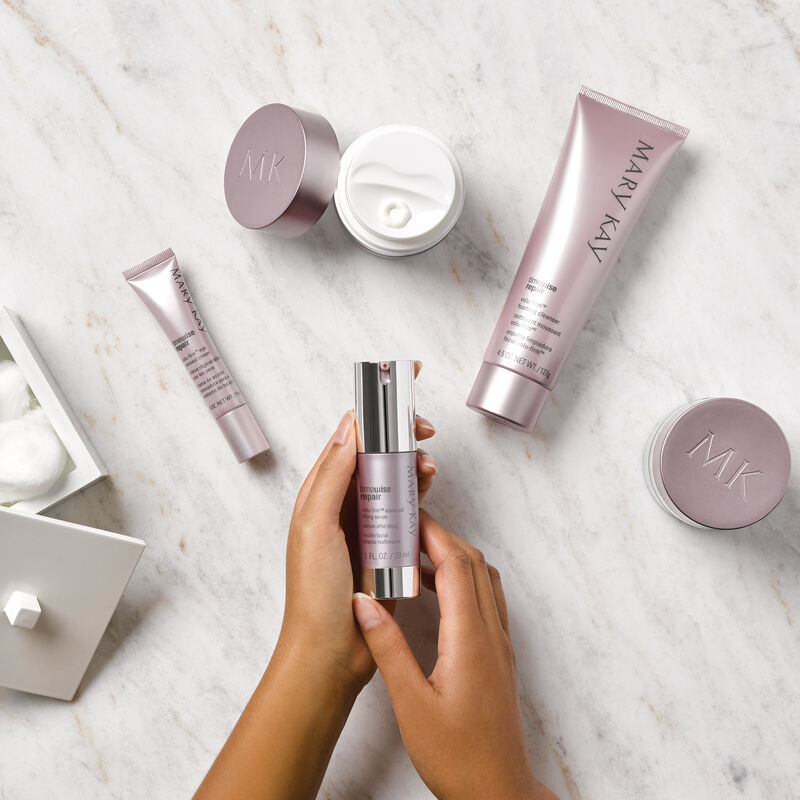 Hands holding TimeWise Repair Volu-Firm Advanced Lifting Serum with other TimeWise Repair products laid on surface
