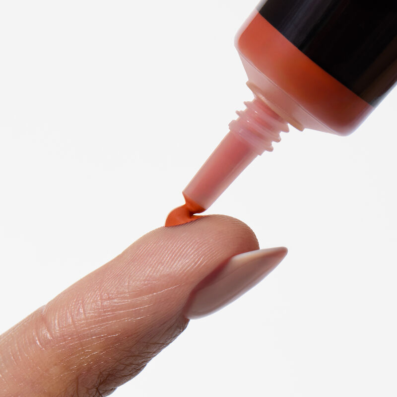 A close-up shot of the Mary Kay Undereye Corrector needle nose squeezing out a dot of product in shade Deep Peach on a finger.